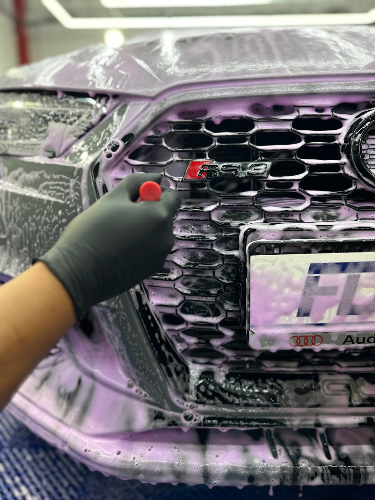 Face avant d’une voiture recouverte de mousse violette, un opérateur en gant noir nettoie la calandre au pinceau